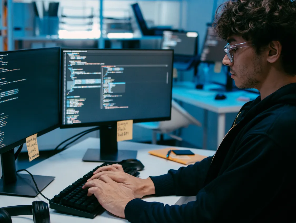 A person in glasses typing at a desk with two monitors of code